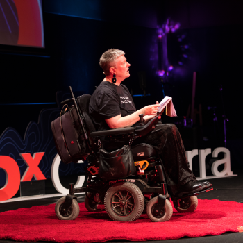 A woman using a power wheelchair presenting on a red-carpeted TEDx Canberra stage under dramatic lighting